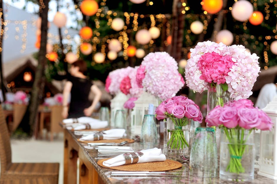 A floral arrangement featuring large, round hydrangea blooms in soft pink and white, paired with smaller bouquets of pink roses in glass vases, displayed on a wooden table set for an event. The flower