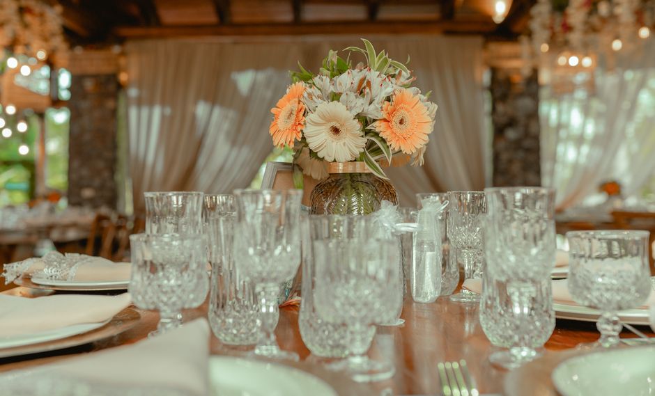 A floral arrangement of light peach and white gerbera daisies, white anemones, and green foliage displayed in a textured brown glass vase, positioned at the center of a wooden dining table. Surroundin