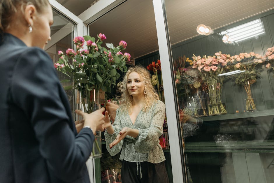 A young woman with curly blonde hair and a floral patterned blouse is standing inside a flower shop, engaged in conversation with a florist who is handing her a bouquet of vibrant pink roses. The bouq