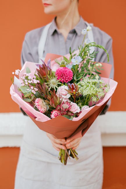 A florist from floristhook.co.uk is holding a vibrant bouquet of fresh flowers arranged with a mix of pink, white, and green blooms, including large pink chrysanthemums, white daisies, and other seaso