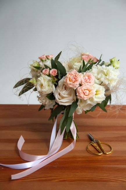 A wedding bouquet consisting of large, soft pink roses, creamy white lisianthus, and delicate blush ranunculus, arranged with dark green eucalyptus leaves and wispy filler flowers. The bouquet is held