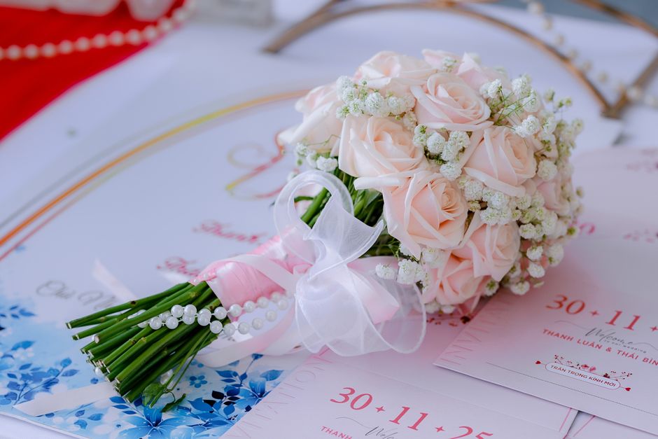 A delicate bouquet featuring soft pink roses and white baby's breath arranged in a compact, round style. The bouquet is wrapped with a light pink satin ribbon tied into a bow, accented with a string o