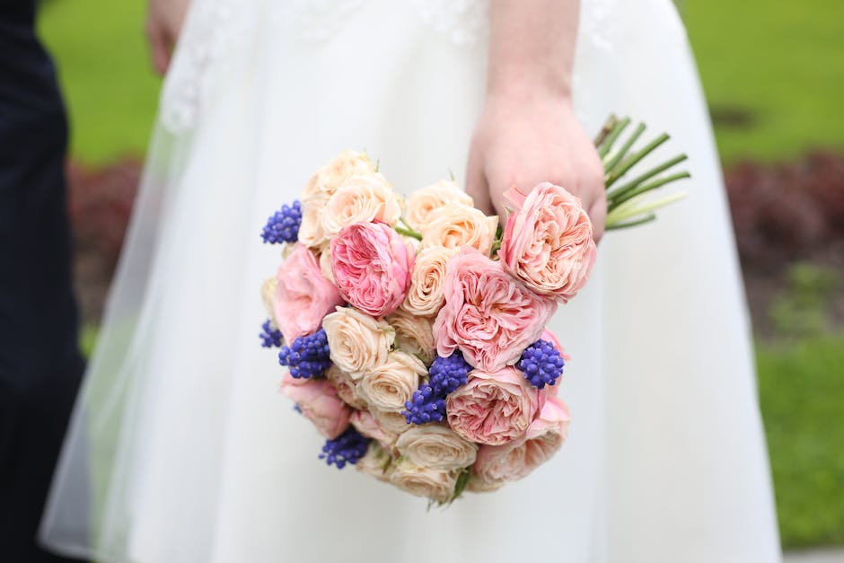 Close-up of a bridal bouquet held by a bride in a white wedding dress, featuring a lush arrangement of pale pink and blush roses, with some deeper pink ranunculus and sprigs of vibrant purple berries 