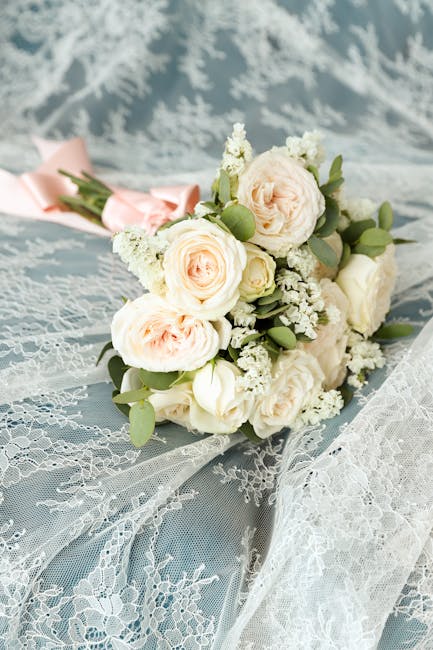 A person dressed in a white lace dress holds a lush floral bouquet featuring white roses, blue hydrangeas, and white garden flowers, accented with green foliage. The bouquet is arranged in a rounded, 
