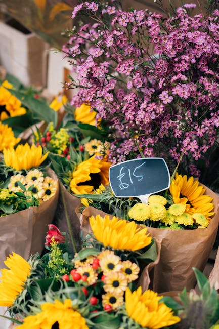 A vibrant assortment of flower bouquets displayed at a market stall, featuring large yellow sunflowers with dark centers, small white and yellow daisies, and bright pink and red blooms, all wrapped in